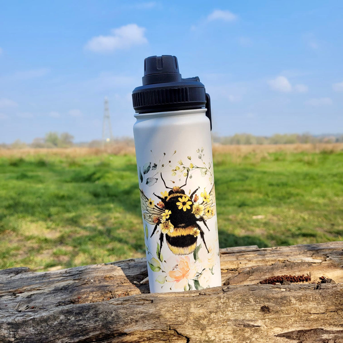 Bee I insulated stainless steel water bottle with a floral bee print, black leak-proof lid, standing on a wooden log.