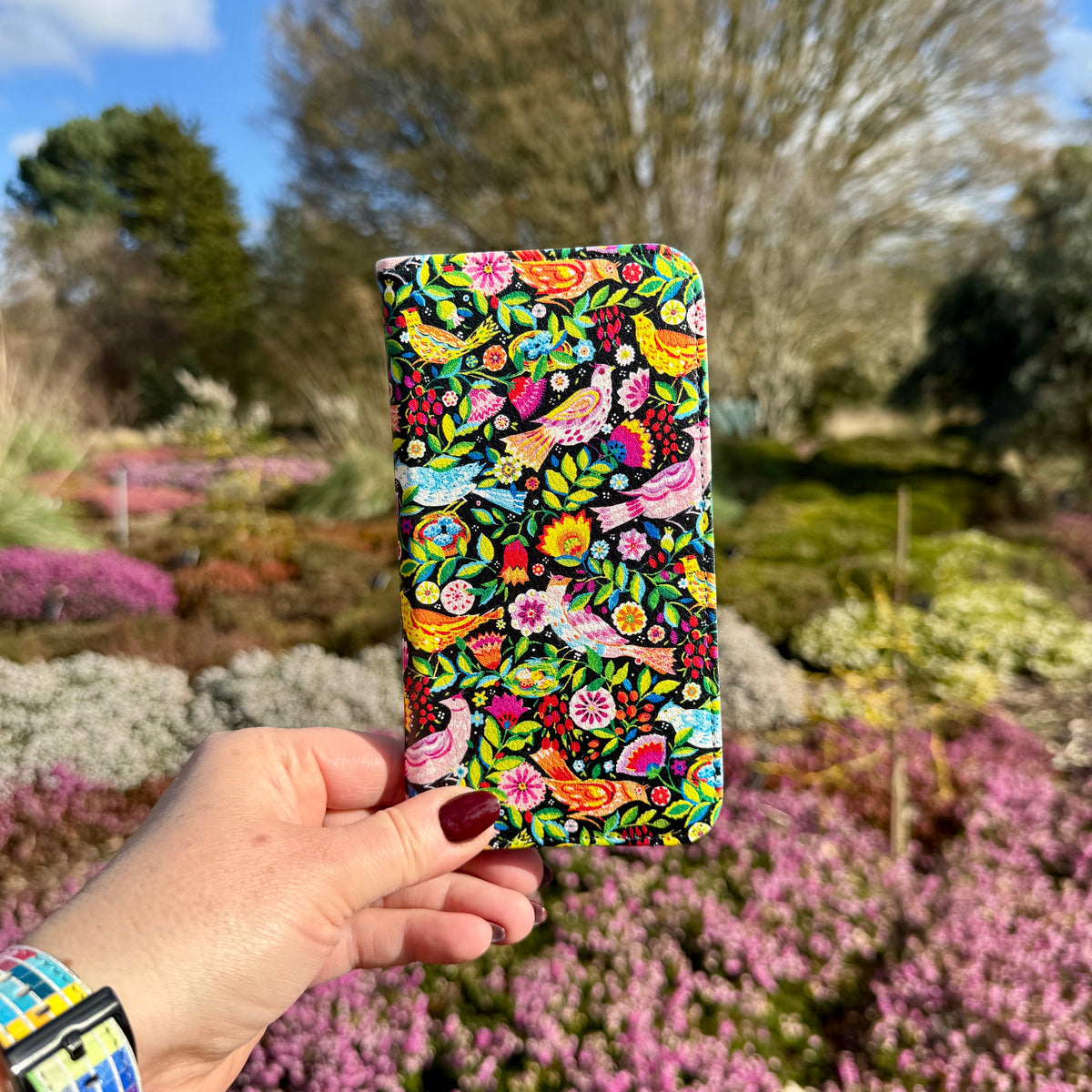 Folkbirds vegan leather folio case with colorful bird and floral print held against a blurred garden background.