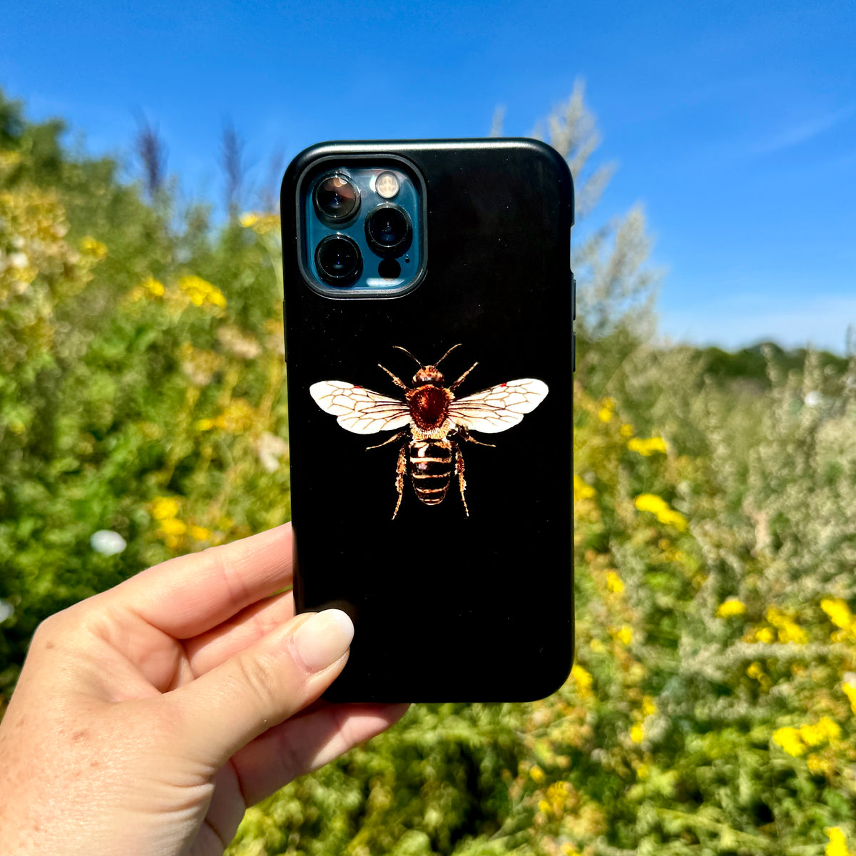Black iPhone tough case featuring a detailed bee illustration, held against a sunny outdoor garden background.