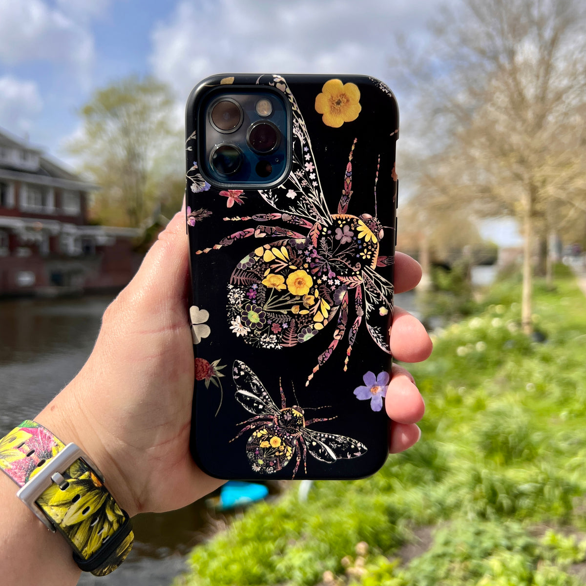 iPhone tough case featuring a black background with intricate floral bumble bee artwork held against a nature backdrop.