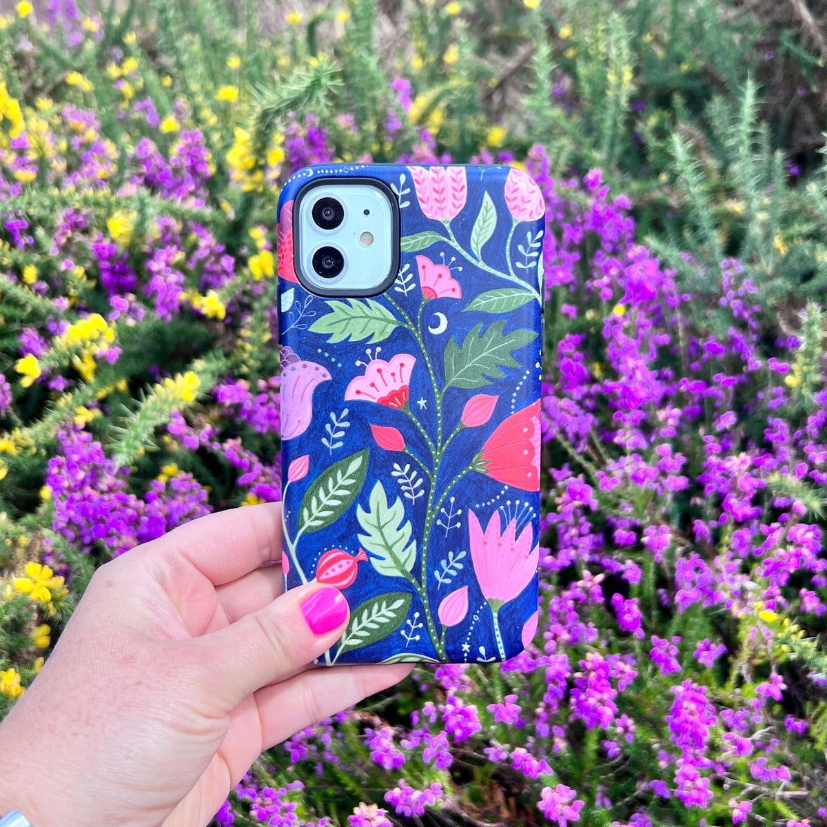Dual-layer iPhone tough case featuring a blue floral pattern by Bex Parkin, held against a backdrop of purple and yellow flowers.