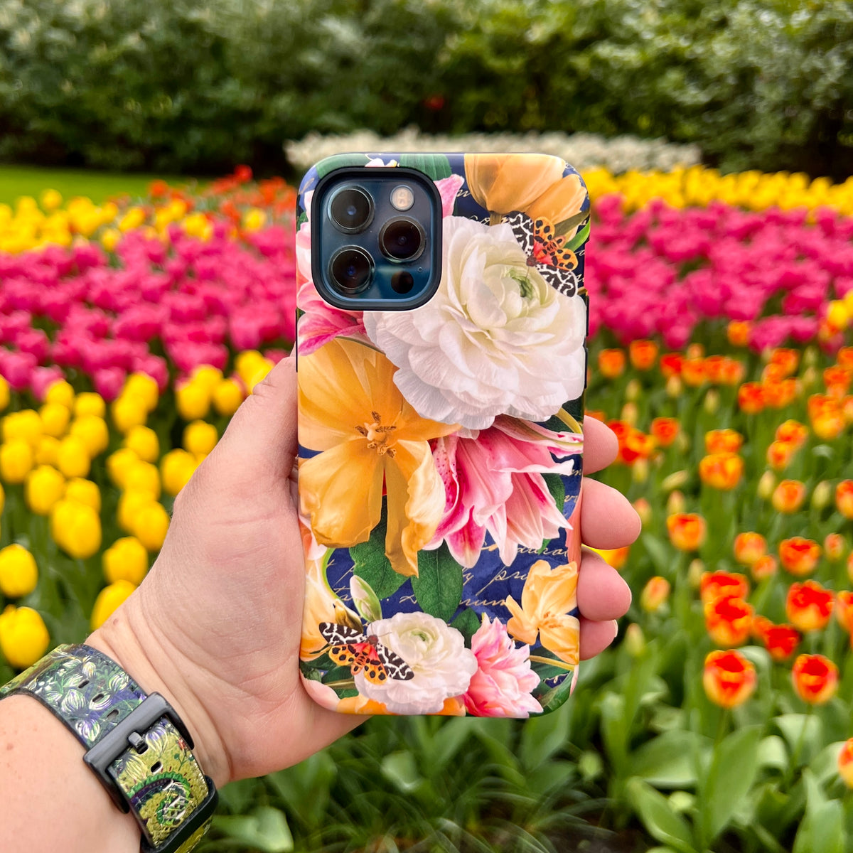 Dual-layer iPhone Tough Case featuring a vibrant floral and butterfly print held against a background of colorful tulips.