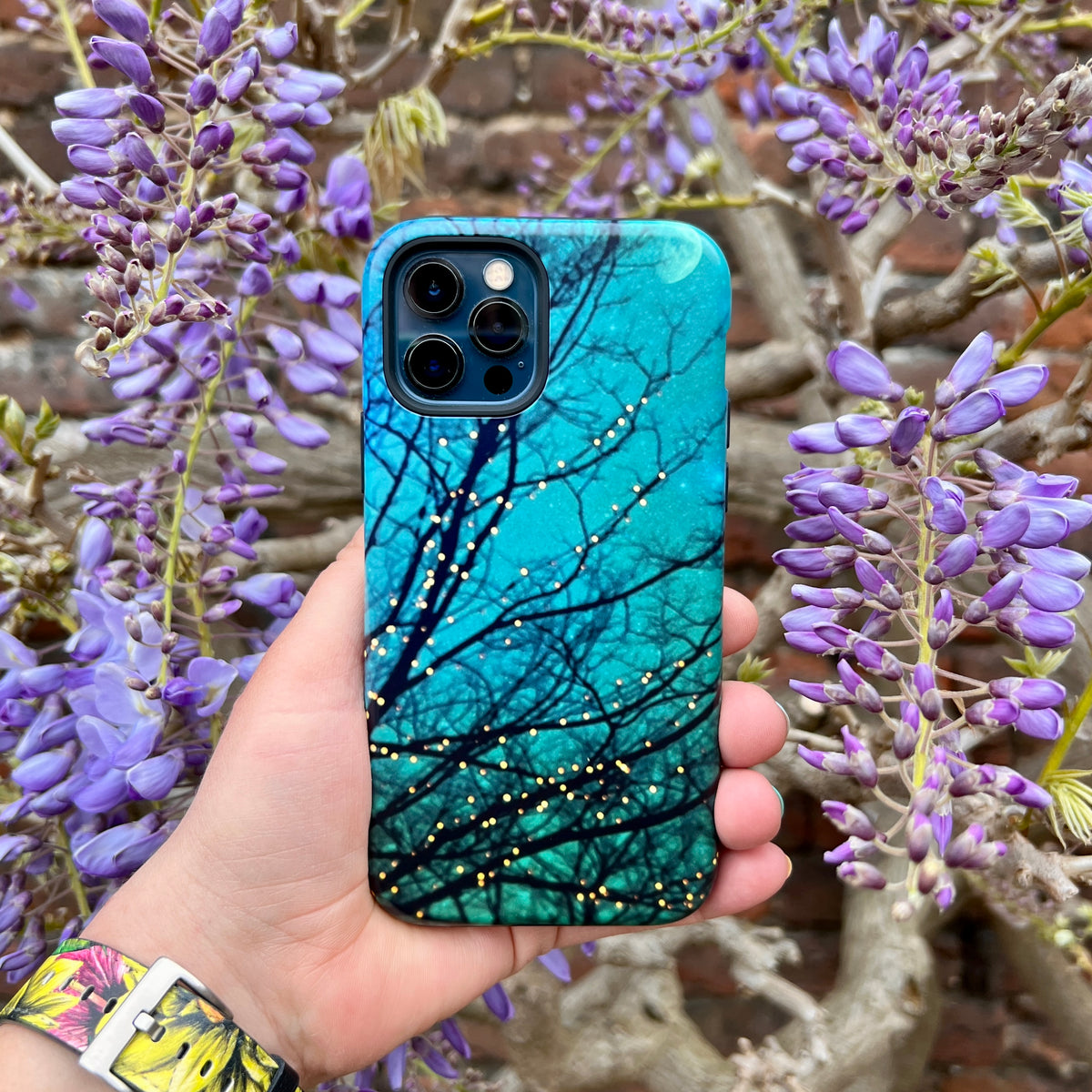 Magical Nights iPhone tough case with teal gradient, dark tree silhouettes, and gold sparkles held against purple wisteria.