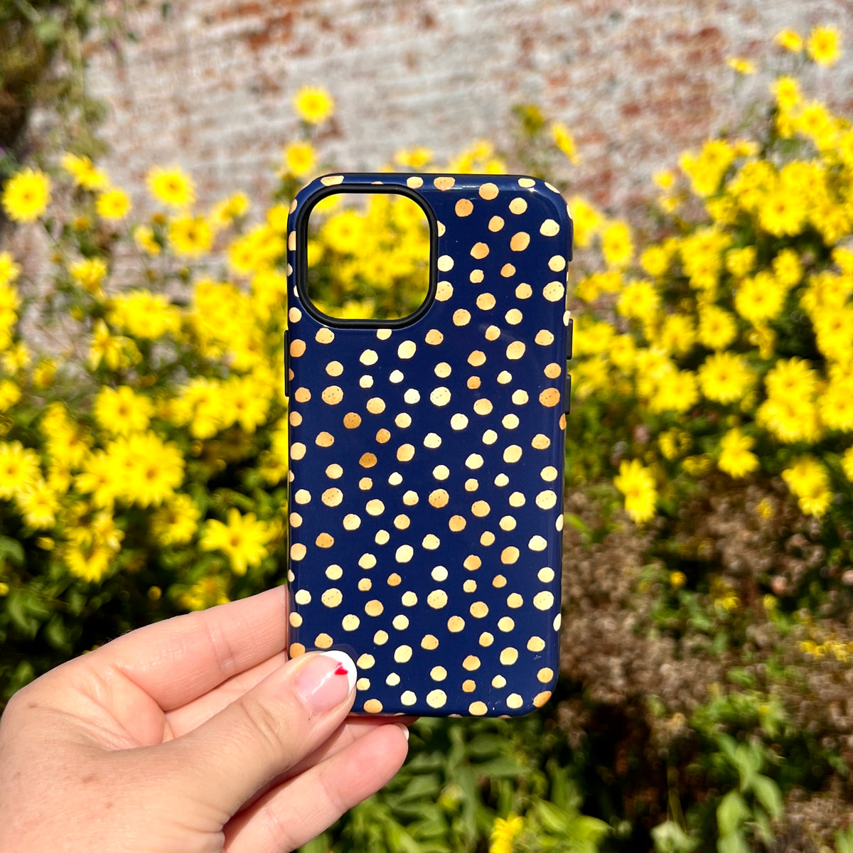 Navy blue iPhone tough case with yellow polka dot pattern, held against a background of yellow flowers.