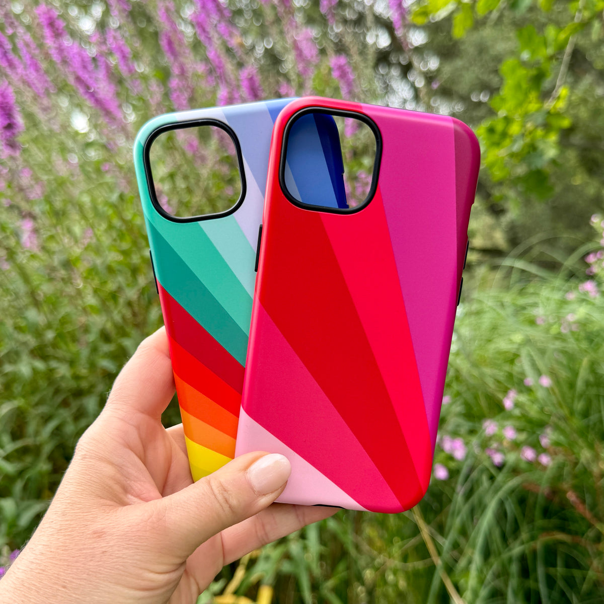 Two dual-layer iPhone tough cases featuring vibrant, multi-colored diagonal stripe patterns held against a garden background.