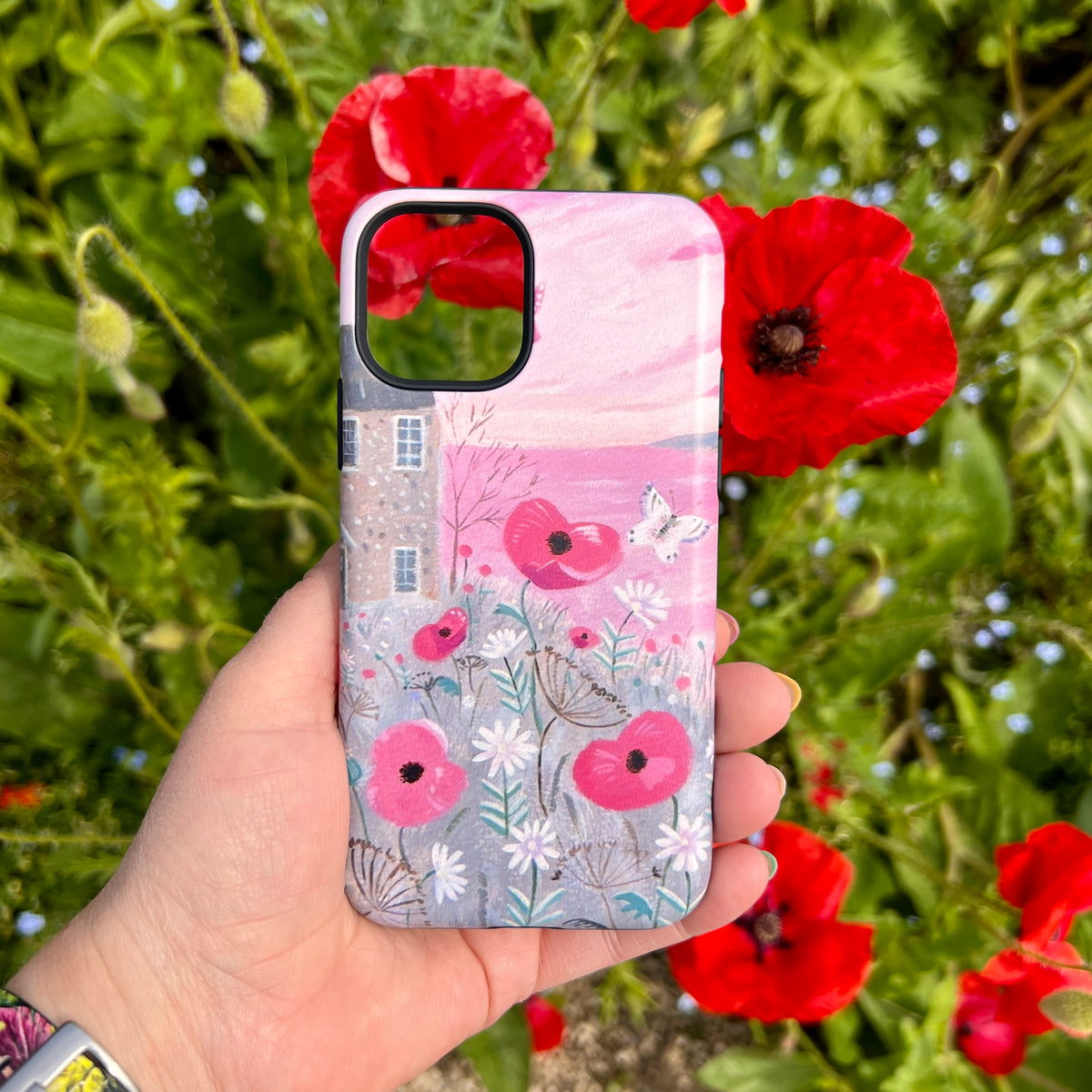 iPhone tough case featuring Seaside Flowers art with a cottage, pink sky, and red poppies held against a garden background.
