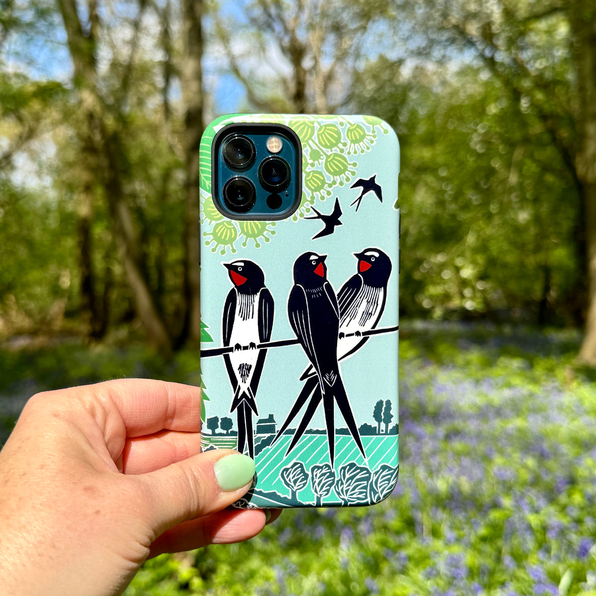 iPhone Tough Case featuring Kate Heiss swallow artwork in blue and green, held against a blurred woodland background.