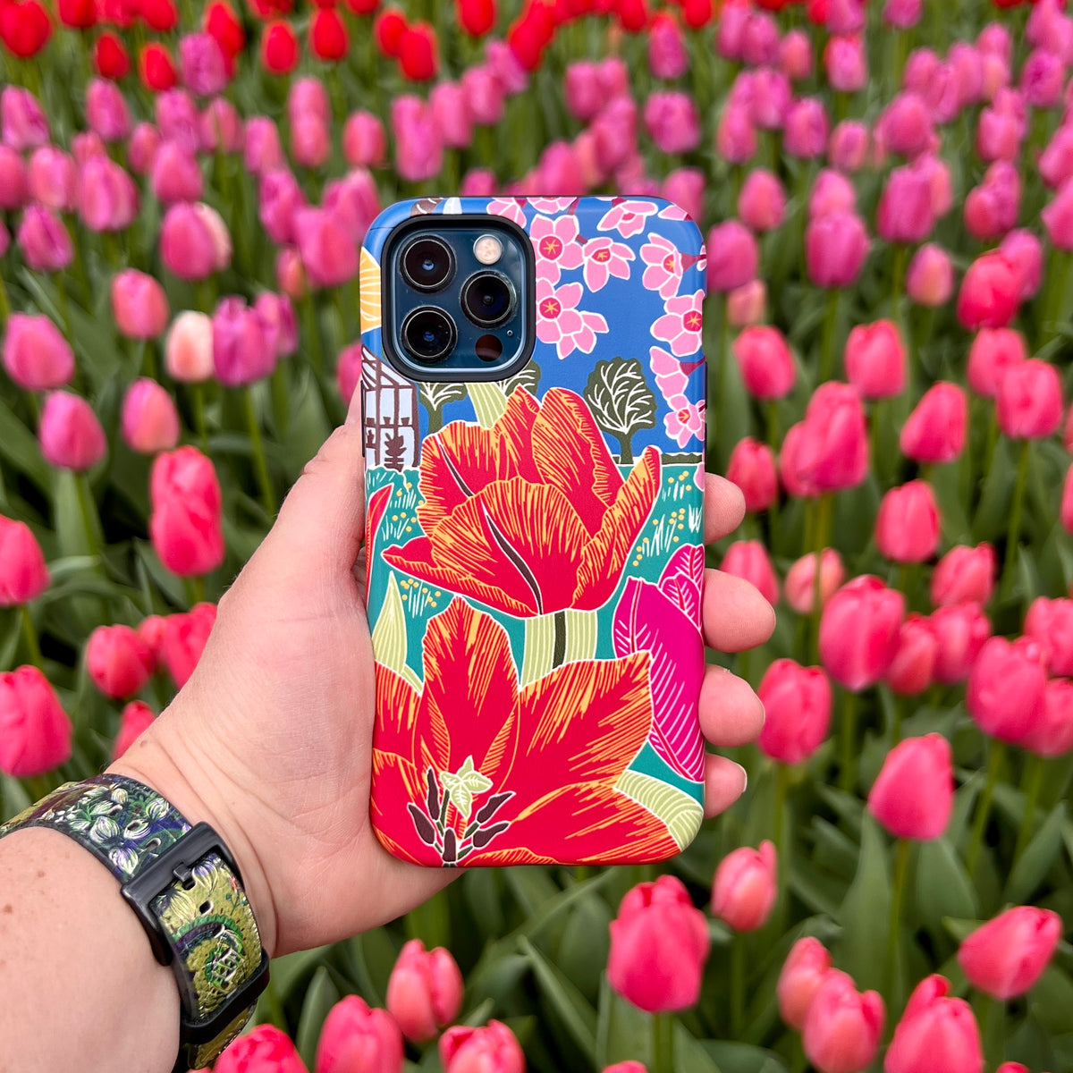 iPhone Tough Case featuring vibrant red tulip artwork by Kate Heiss, held against a background of blooming pink tulips.