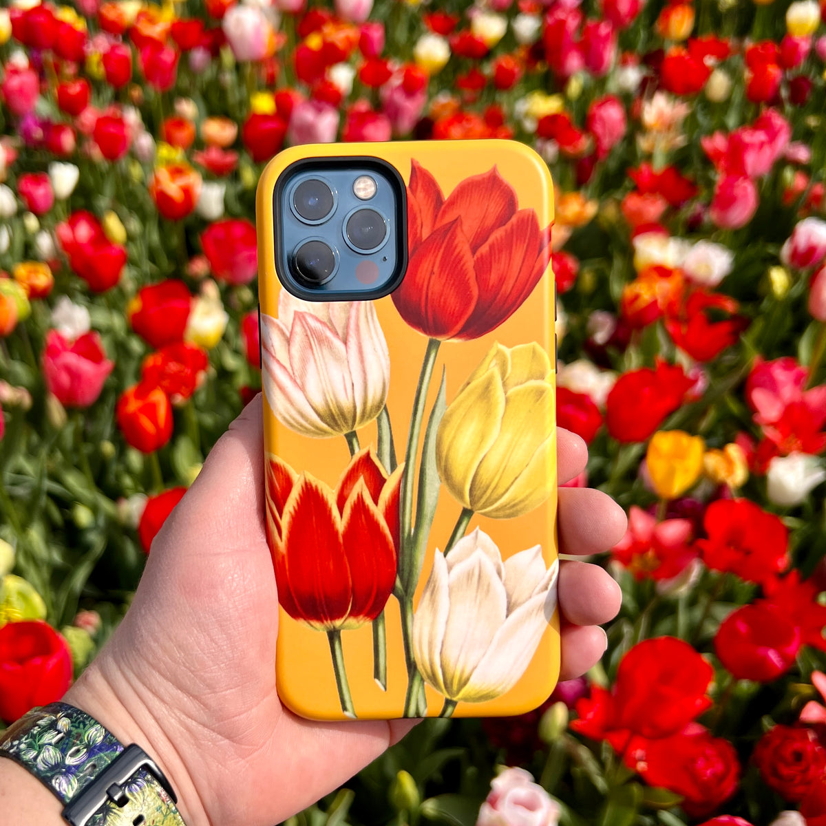 Hand holding a yellow iPhone tough case featuring a vibrant red, yellow, and white tulip floral print against a tulip field.