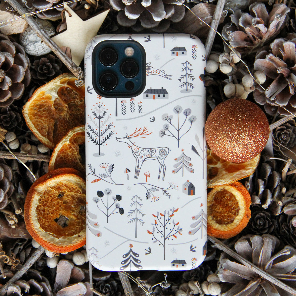 White iPhone tough case featuring a Winterfold deer and forest pattern, surrounded by pinecones and dried orange slices.