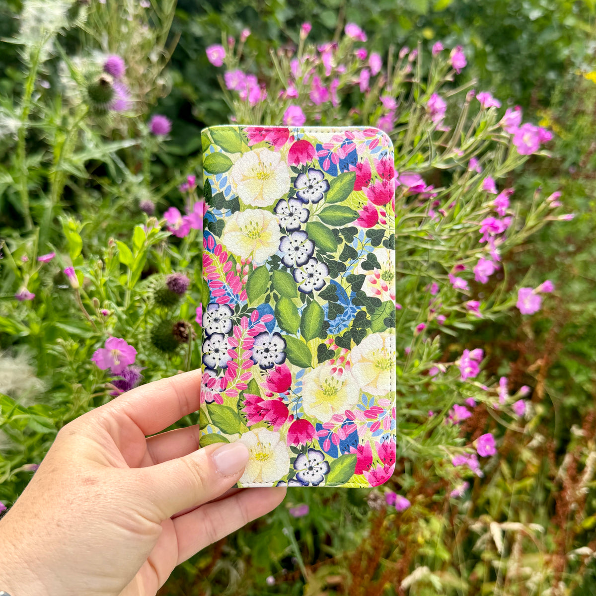 Vegan leather folio phone case with vibrant floral artwork held against a background of pink garden flowers.