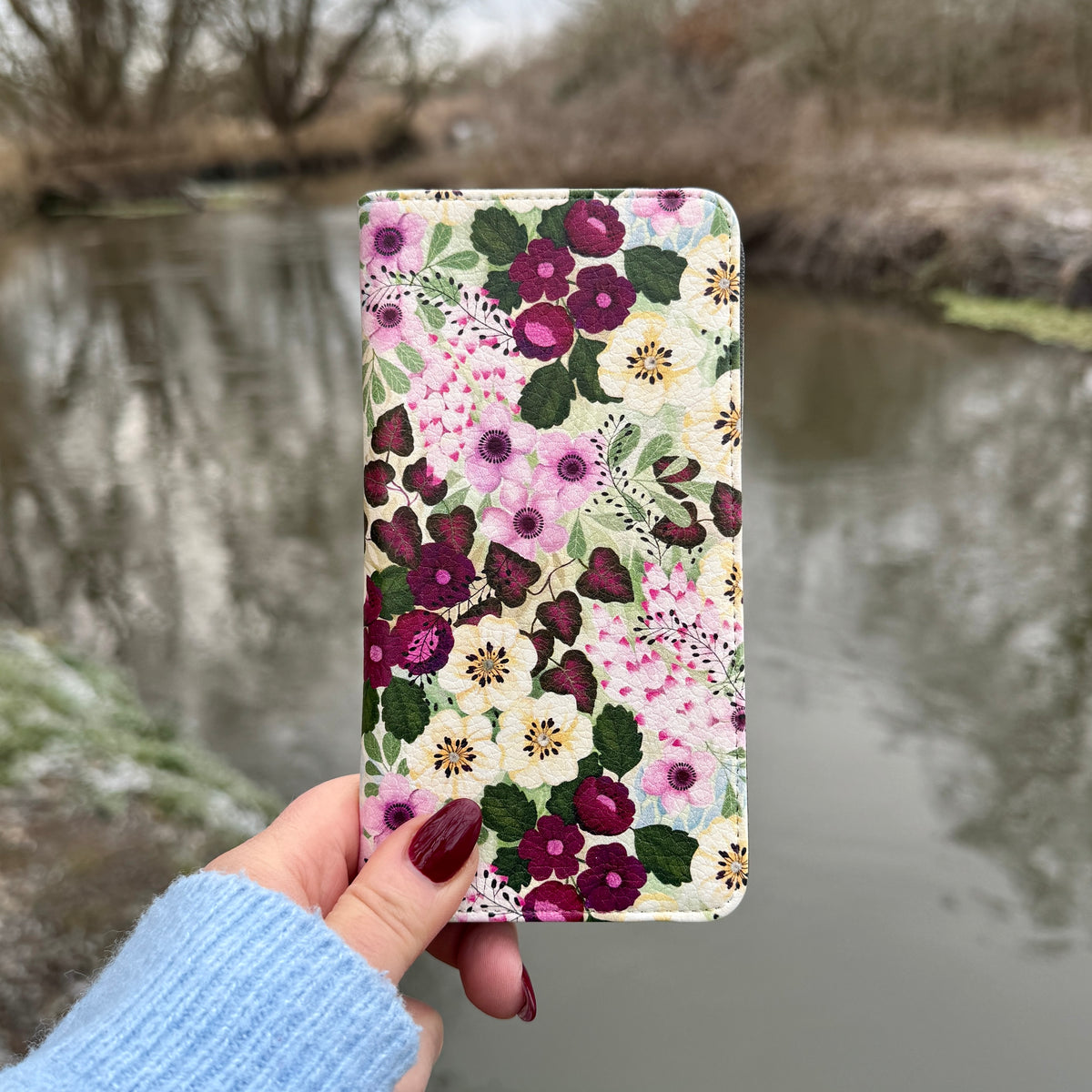 Purple floral vegan leather folio phone case with a satin finish held against a blurred river background.