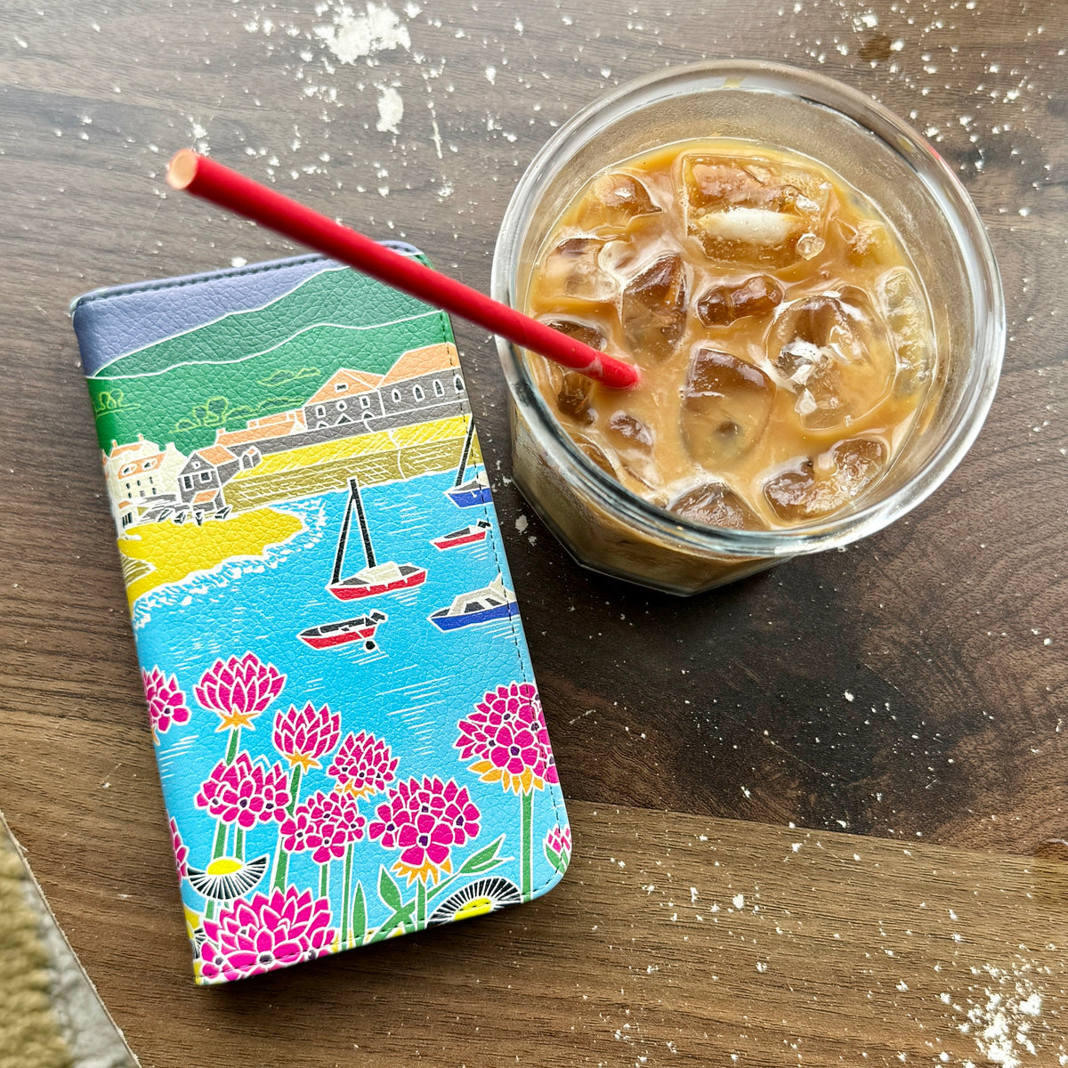 Quaint Harbour vegan leather folio phone case with colorful coastal artwork next to an iced coffee on a wooden table.