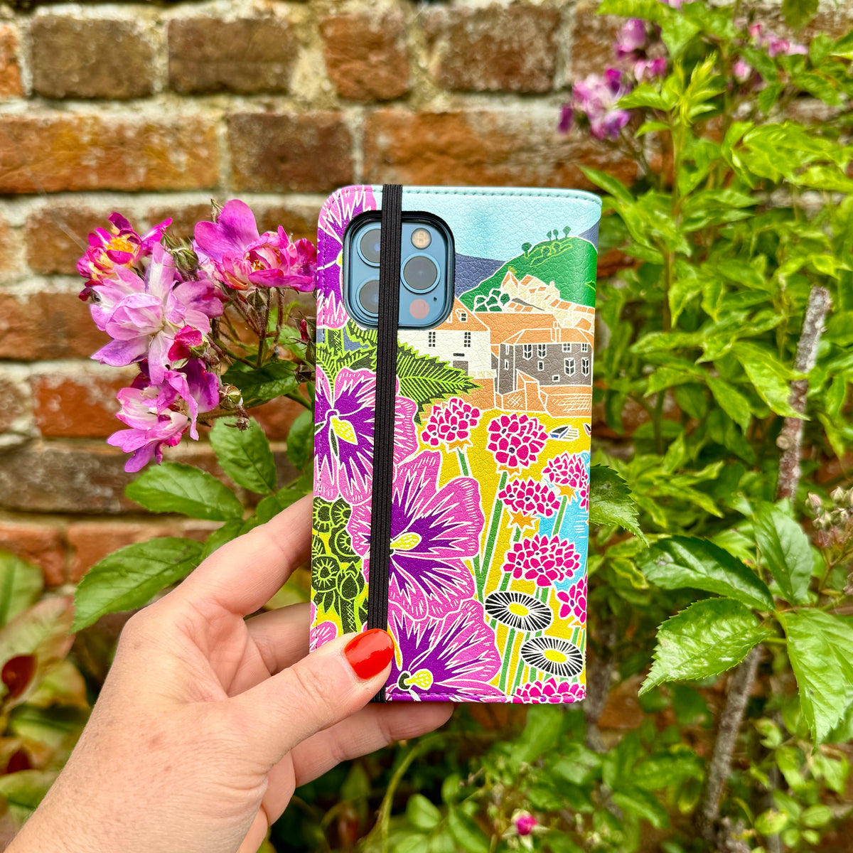 Vegan leather phone folio case featuring Kate Heiss Quaint Harbour artwork with vibrant floral prints and a black elastic strap.