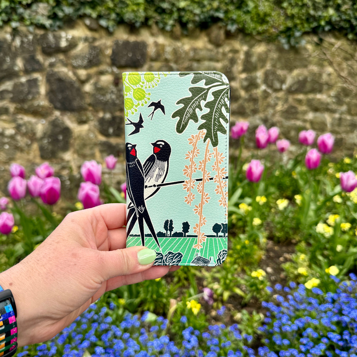 Vegan leather folio phone case featuring Kate Heiss swallow artwork in a light blue satin finish held in a garden.