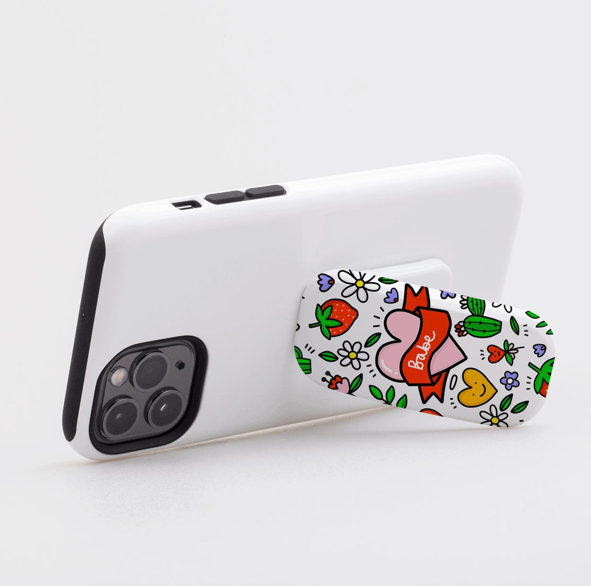 Babe phone stand with a colorful heart, strawberry, and floral pattern attached to the back of a white smartphone.