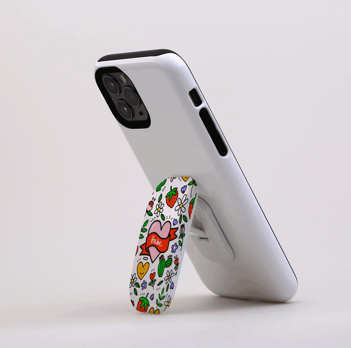 White smartphone propped up by a Flickit stand featuring a colorful heart, strawberry, and floral pattern.