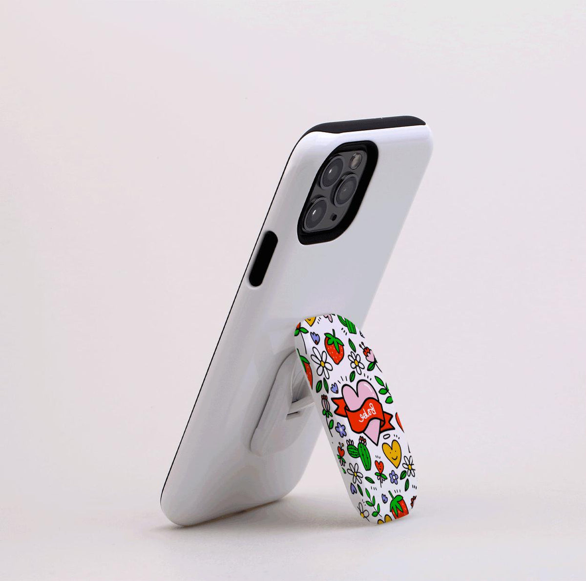 White smartphone propped up by a Babe Flickit stand featuring a colorful strawberry, heart, and floral pattern.
