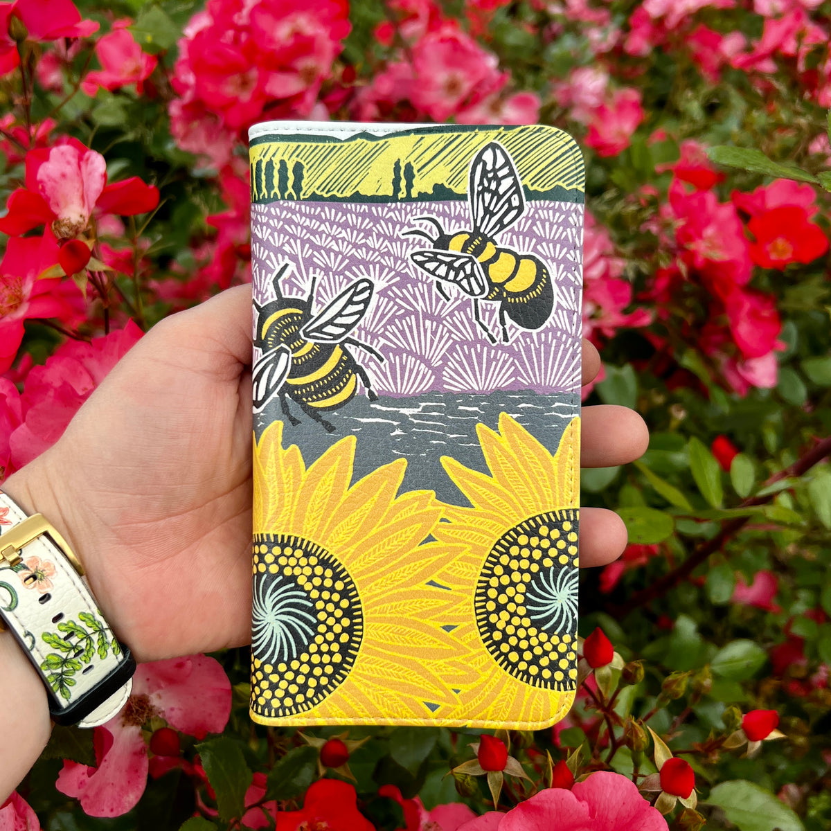 Vegan leather folio phone case featuring Kate Heiss bee and sunflower art with a satin finish held against pink roses.