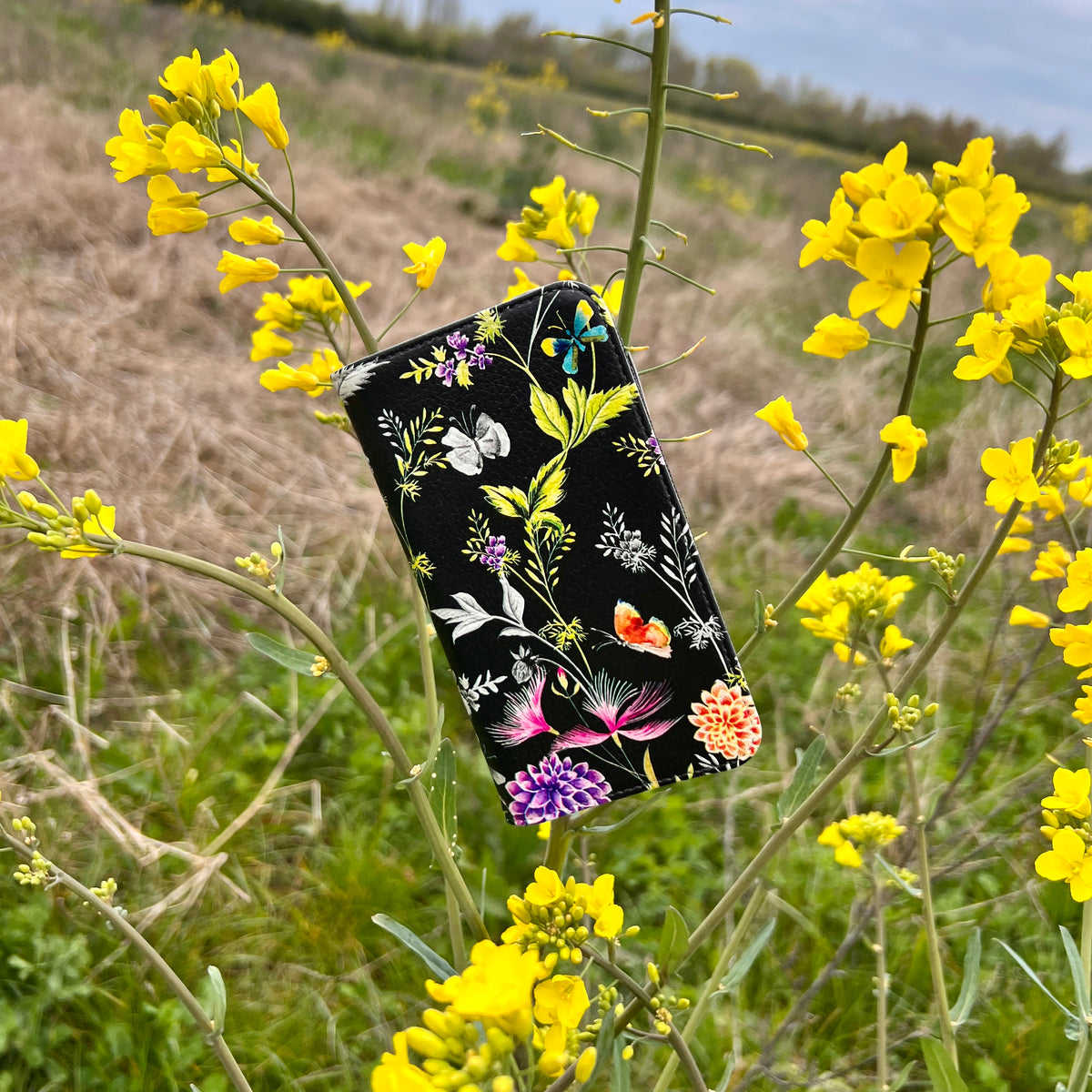 Broadview Gardens vegan leather folio phone case with a black floral and butterfly print, displayed among yellow wildflowers.