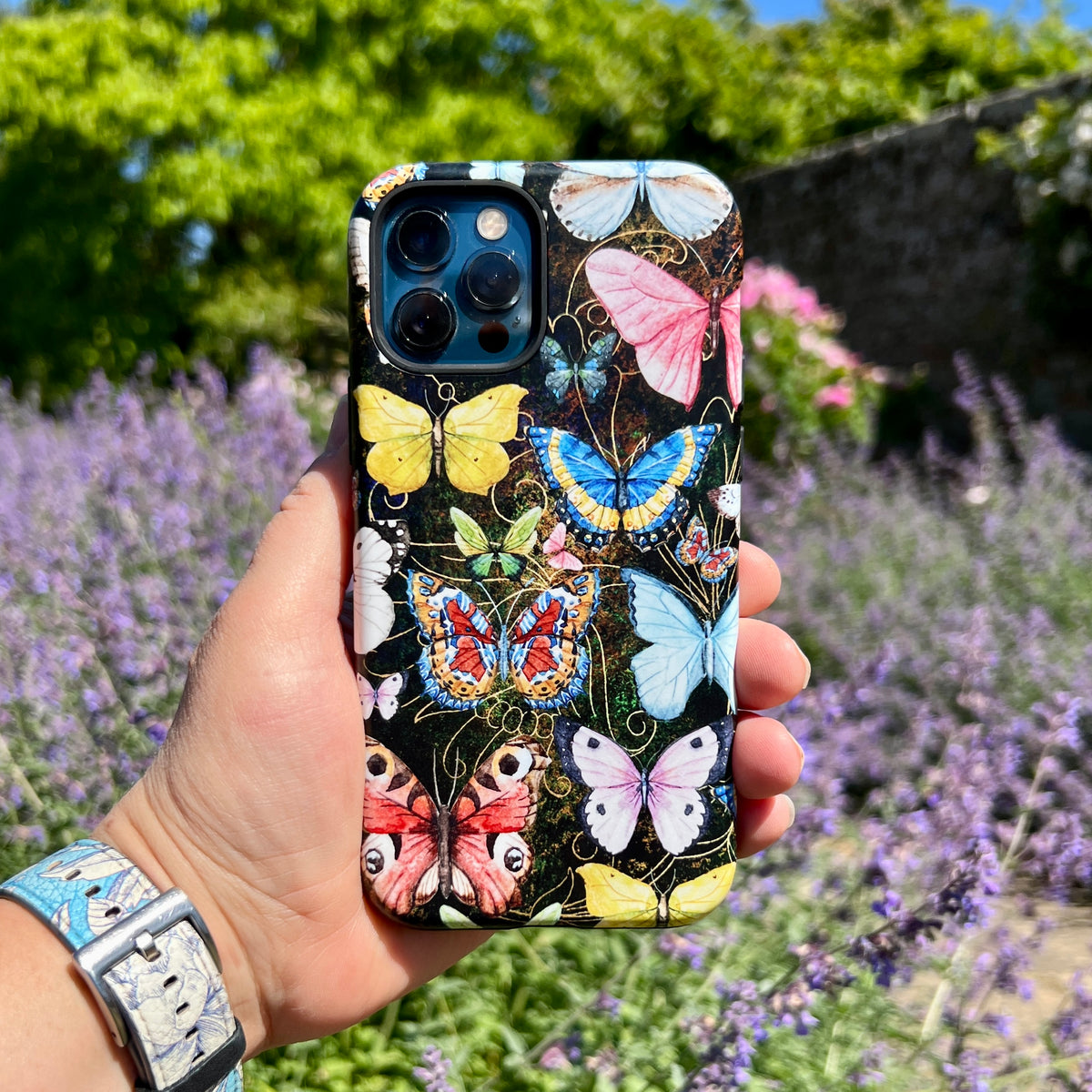 iPhone Tough Case featuring a vibrant, multi-colored butterfly pattern on a dark background held against a garden backdrop.