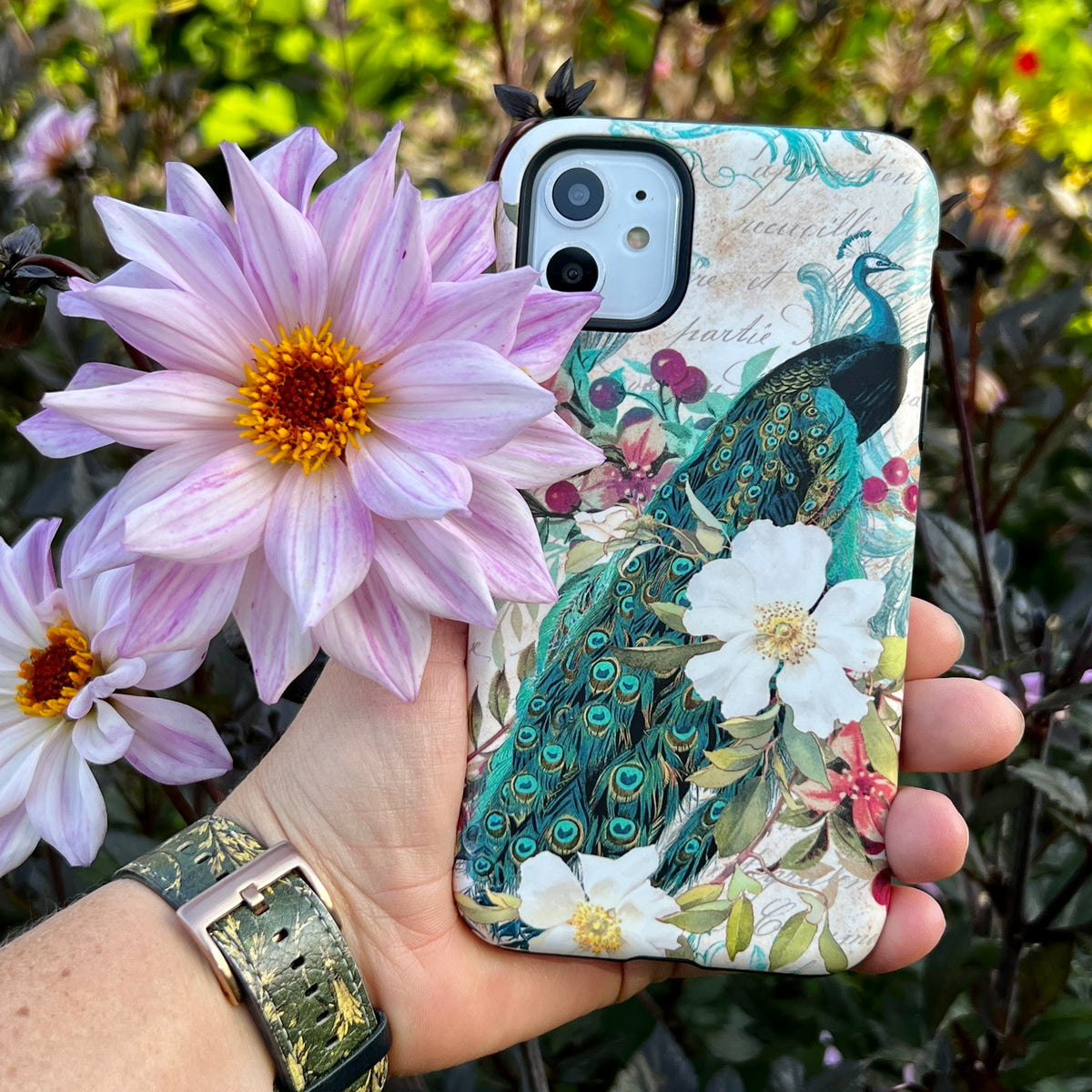 Dual-layer iPhone tough case featuring a vibrant peacock and floral art design, held in hand against a garden background.