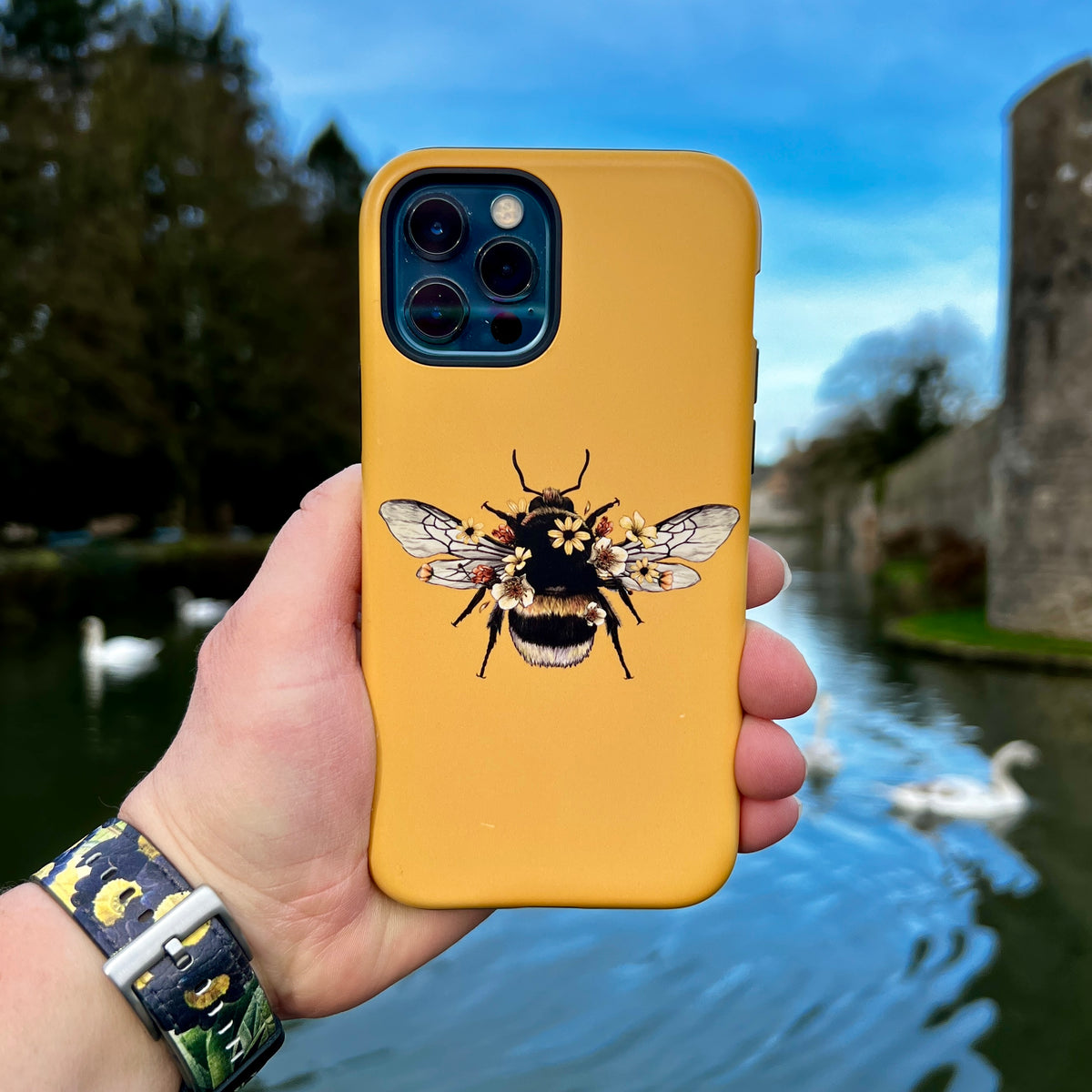Mustard yellow iPhone tough case featuring a detailed bumblebee and floral illustration held against a river background.