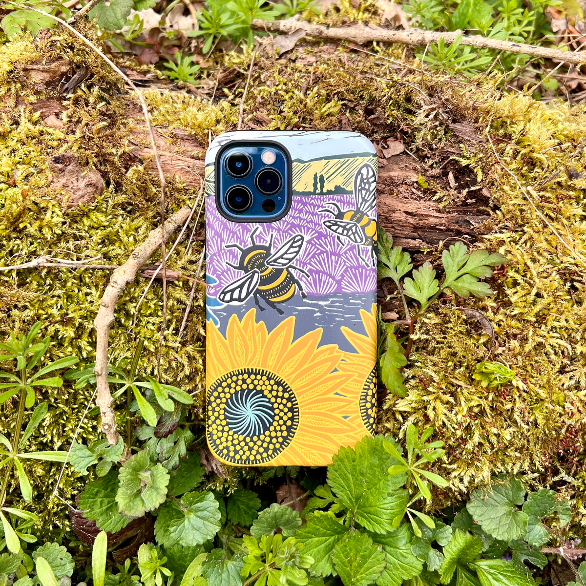 iPhone tough case featuring Kate Heiss bee and sunflower art, resting on mossy ground with dual-layer protective design.