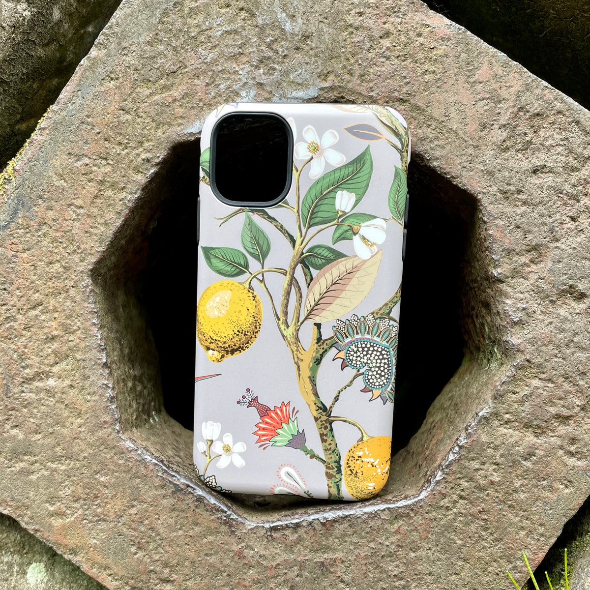 iPhone Tough Case featuring a botanical Burnby Hall print with lemons and flowers on a grey background, resting on stone.