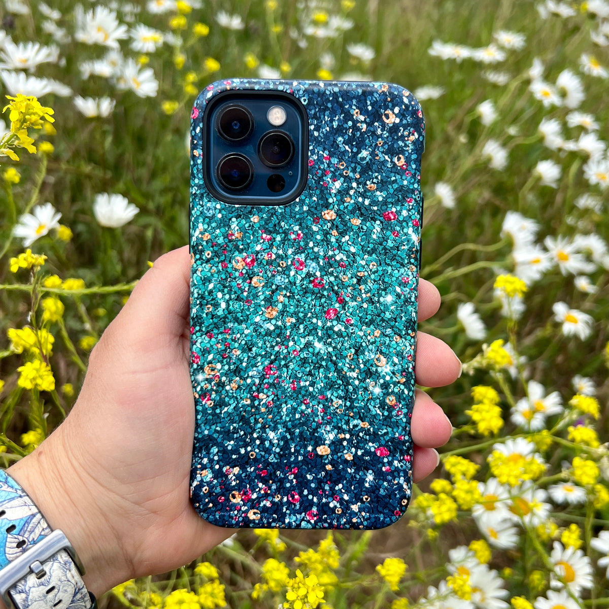 Butterfly Comet dual-layer iPhone tough case with a teal and multicolored printed pattern held against a field of flowers.