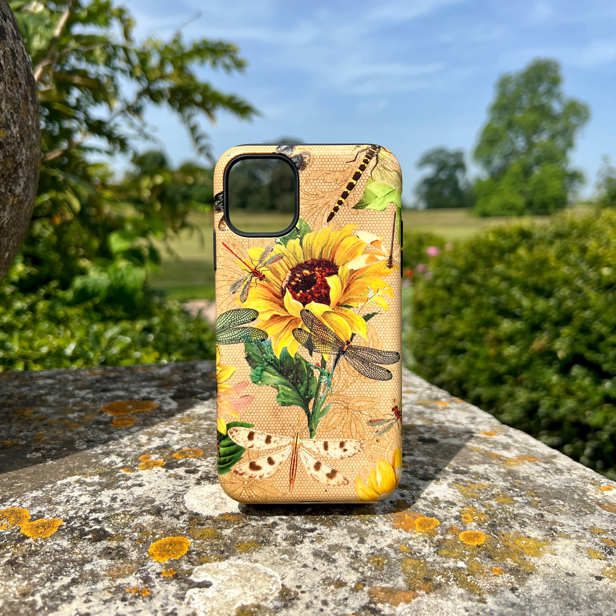 iPhone tough case with a dragonfly and sunflower pattern on a honeycomb background, resting on a stone surface outdoors.