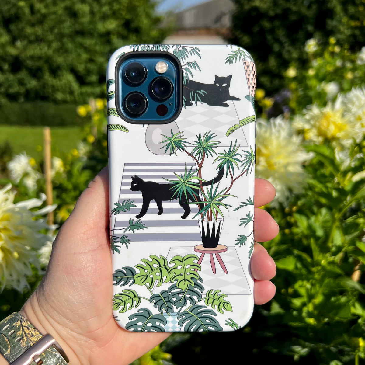 Flimby iPhone tough case with a black cat and houseplant pattern held against a blurred garden background.
