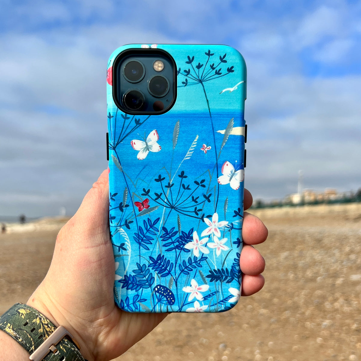 iPhone tough case featuring blue floral and butterfly artwork by Mary Stubberfield, held against a beach background.