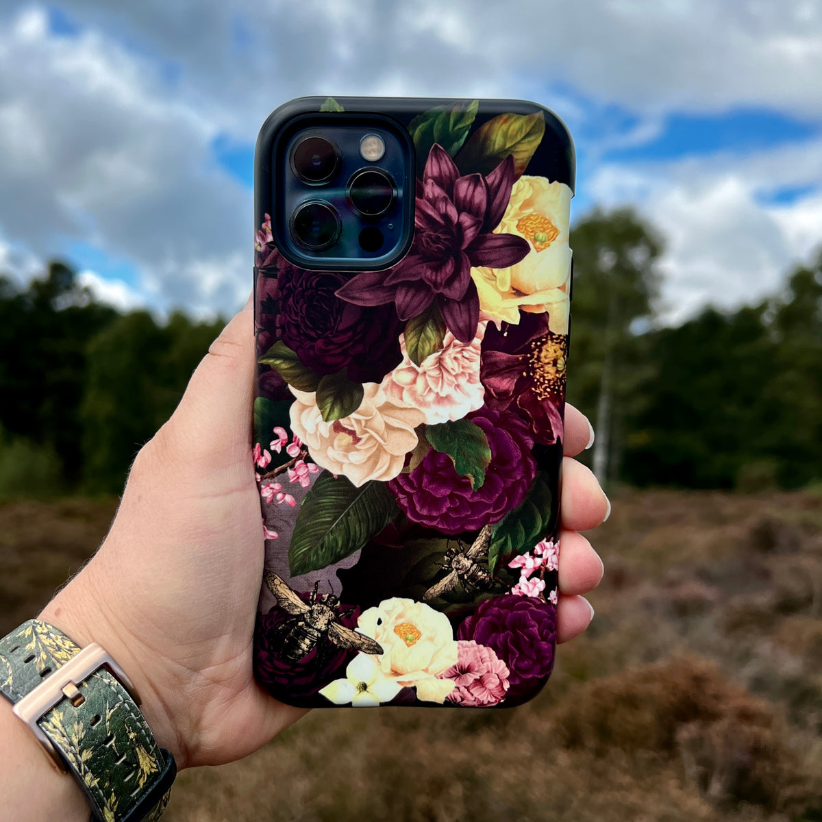 iPhone Tough Case featuring a dark floral design with bees, held in hand against an outdoor landscape background.