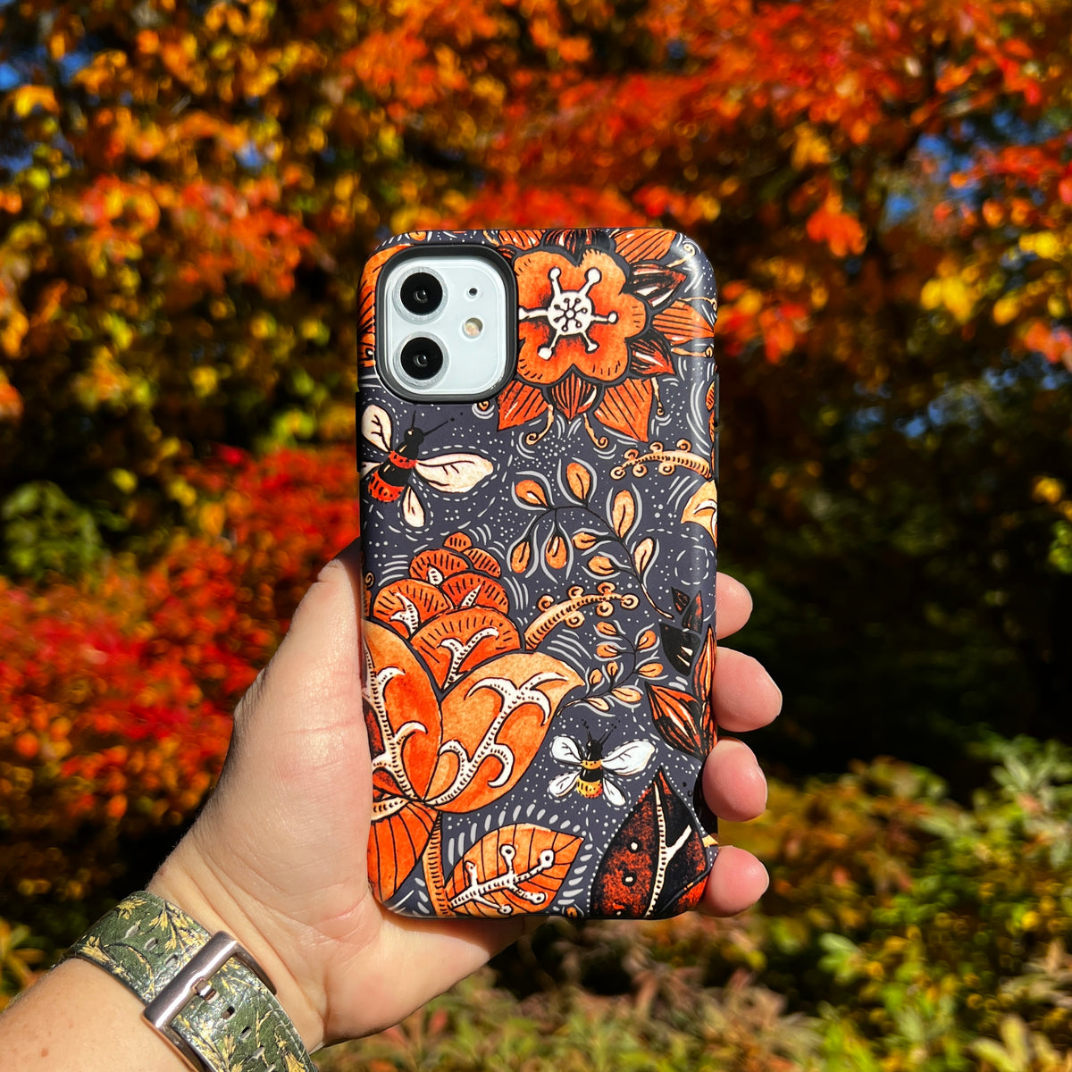 iPhone Tough Case in Hidcote design featuring orange floral and bee patterns on a dark grey background held outdoors.