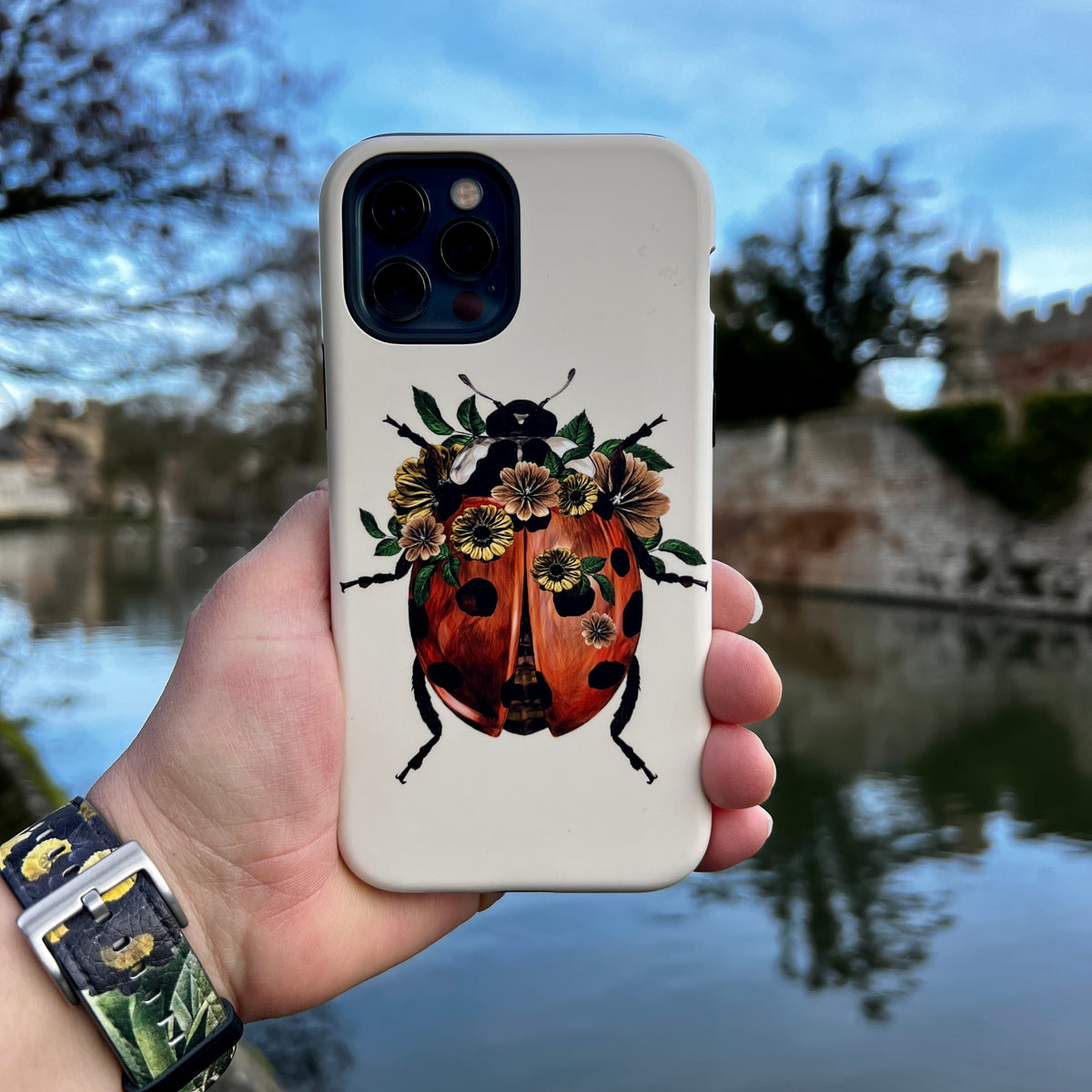 Off-white iPhone tough case featuring a detailed ladybug illustration adorned with small flowers, held against a river.