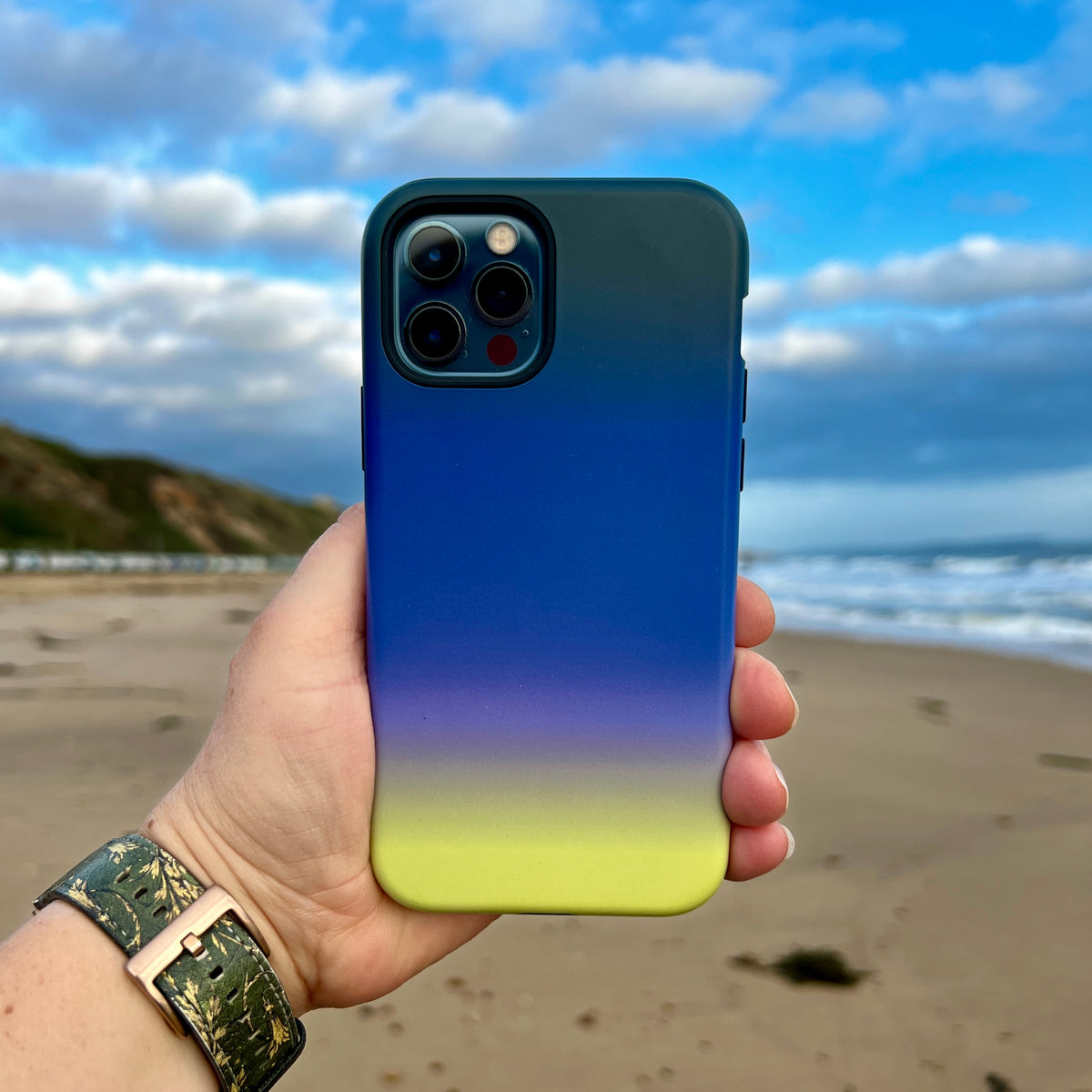Lavender Haze iPhone tough case with a smooth gradient from dark green to blue, purple, and yellow held on a beach.