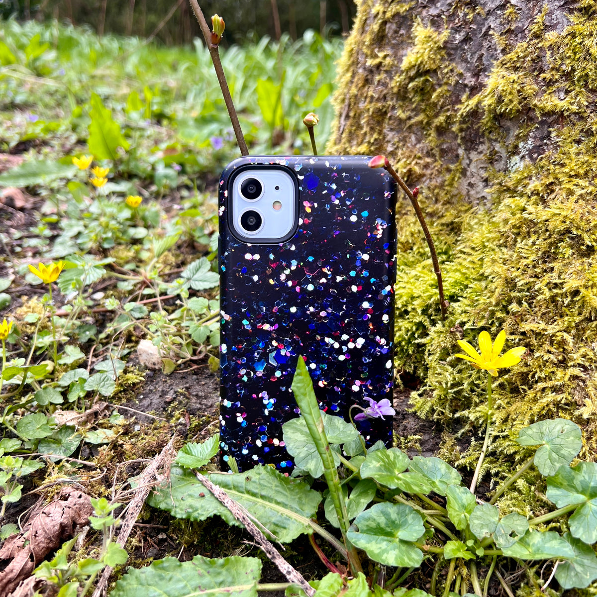 iPhone tough case with a dark Night Sky print featuring colorful abstract spots, resting against a mossy tree in a garden.
