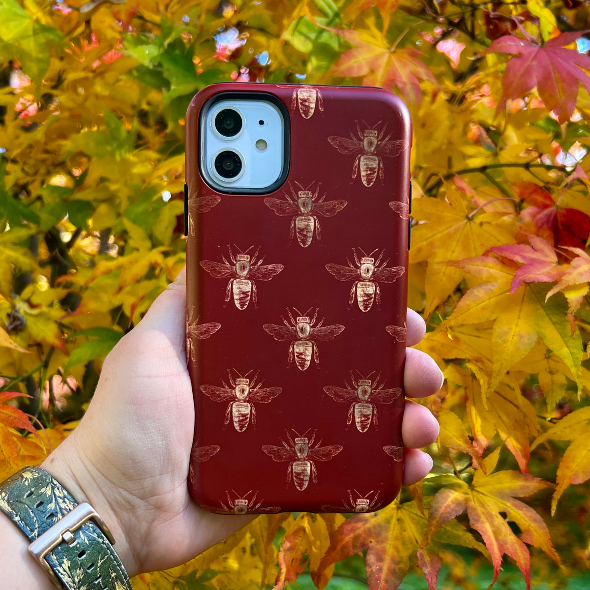 Red iPhone tough case with a gold bee pattern held in hand against a backdrop of vibrant autumn maple leaves.