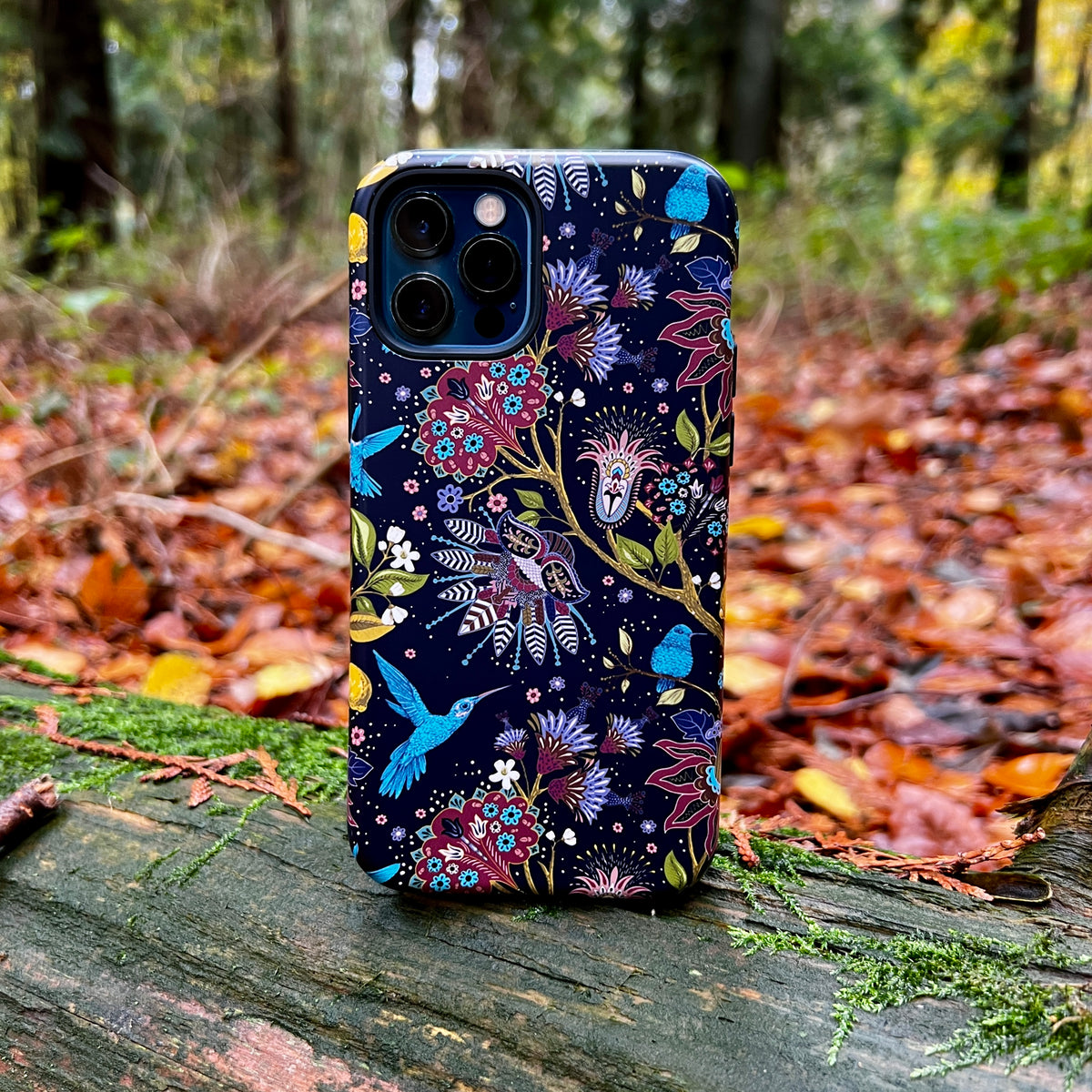 iPhone Tough Case with a dark floral and hummingbird pattern resting on a mossy log in an autumn forest.