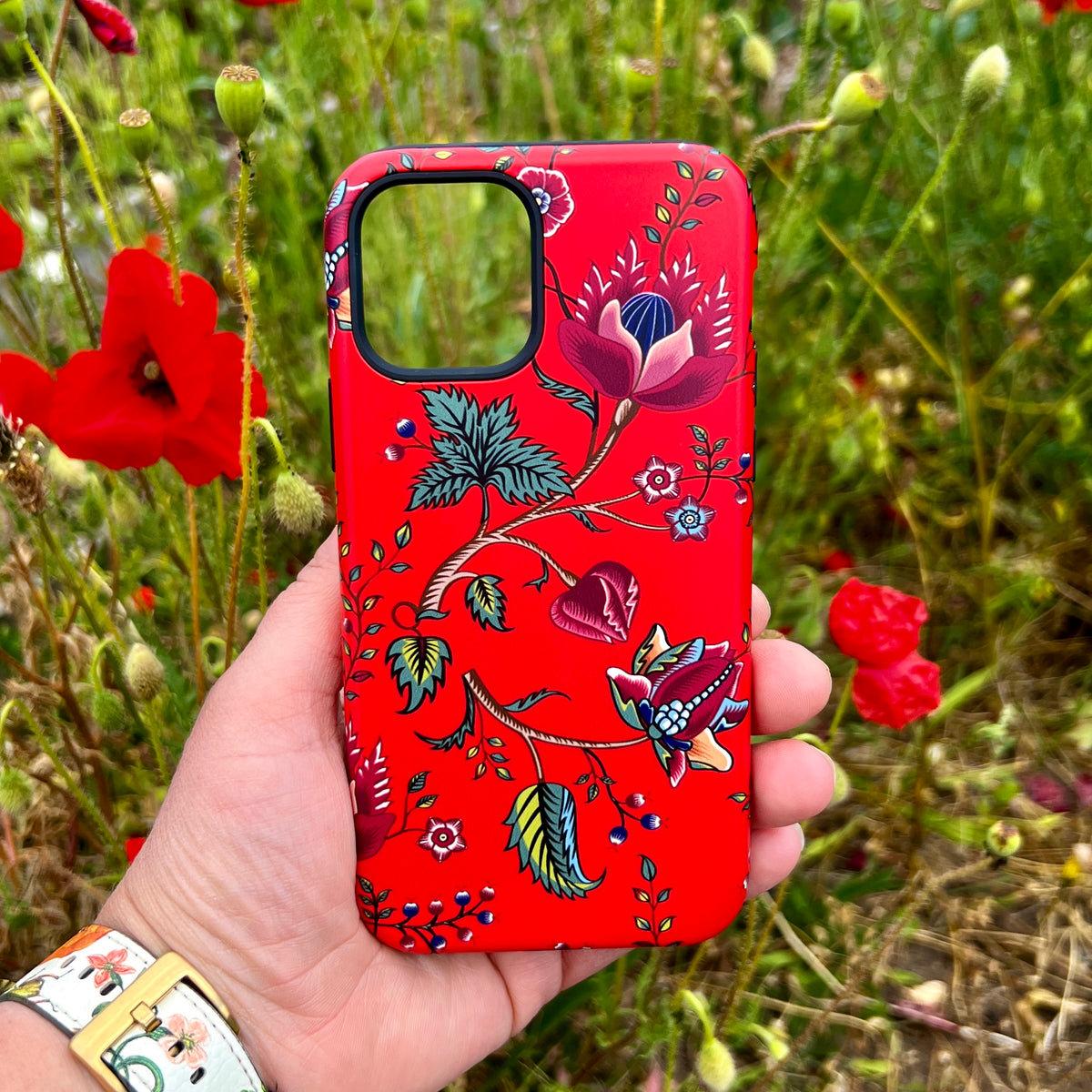 Red iPhone tough case featuring a vibrant floral botanical print held against a background of blooming red poppy flowers.