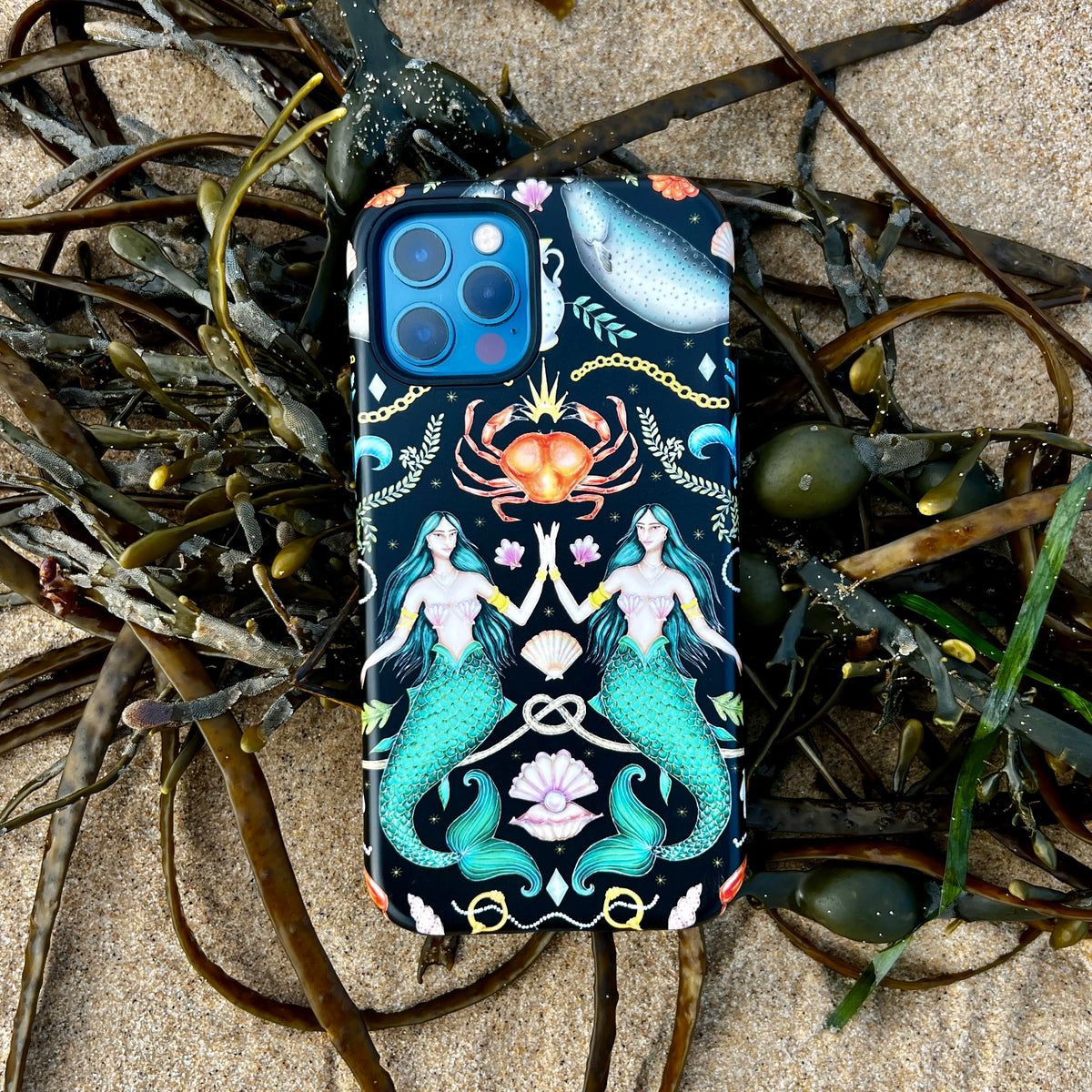 Tritons Treasure iPhone tough case featuring mermaid and crab art on a black background, resting on sandy beach seaweed.