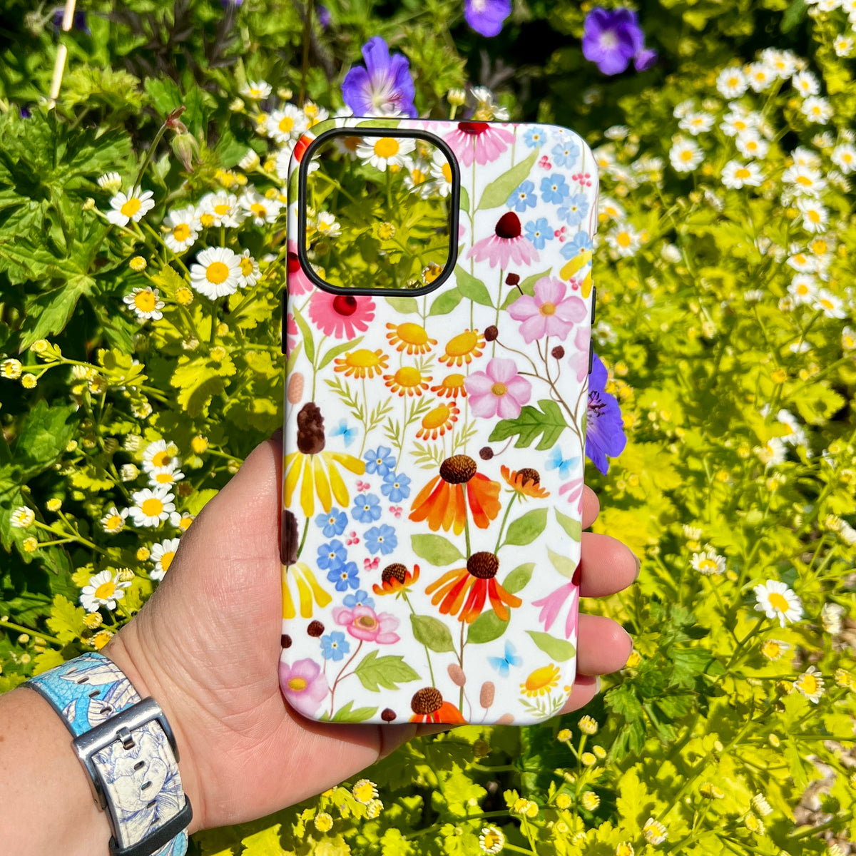 iPhone tough case with a colorful wildflower pattern held against a background of blooming garden flowers.