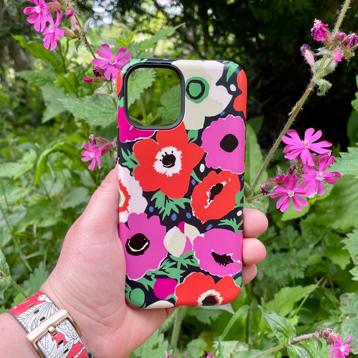 iPhone Tough Case featuring Sarah Campbell’s vibrant red and pink Windflowers floral print against a lush garden backdrop.