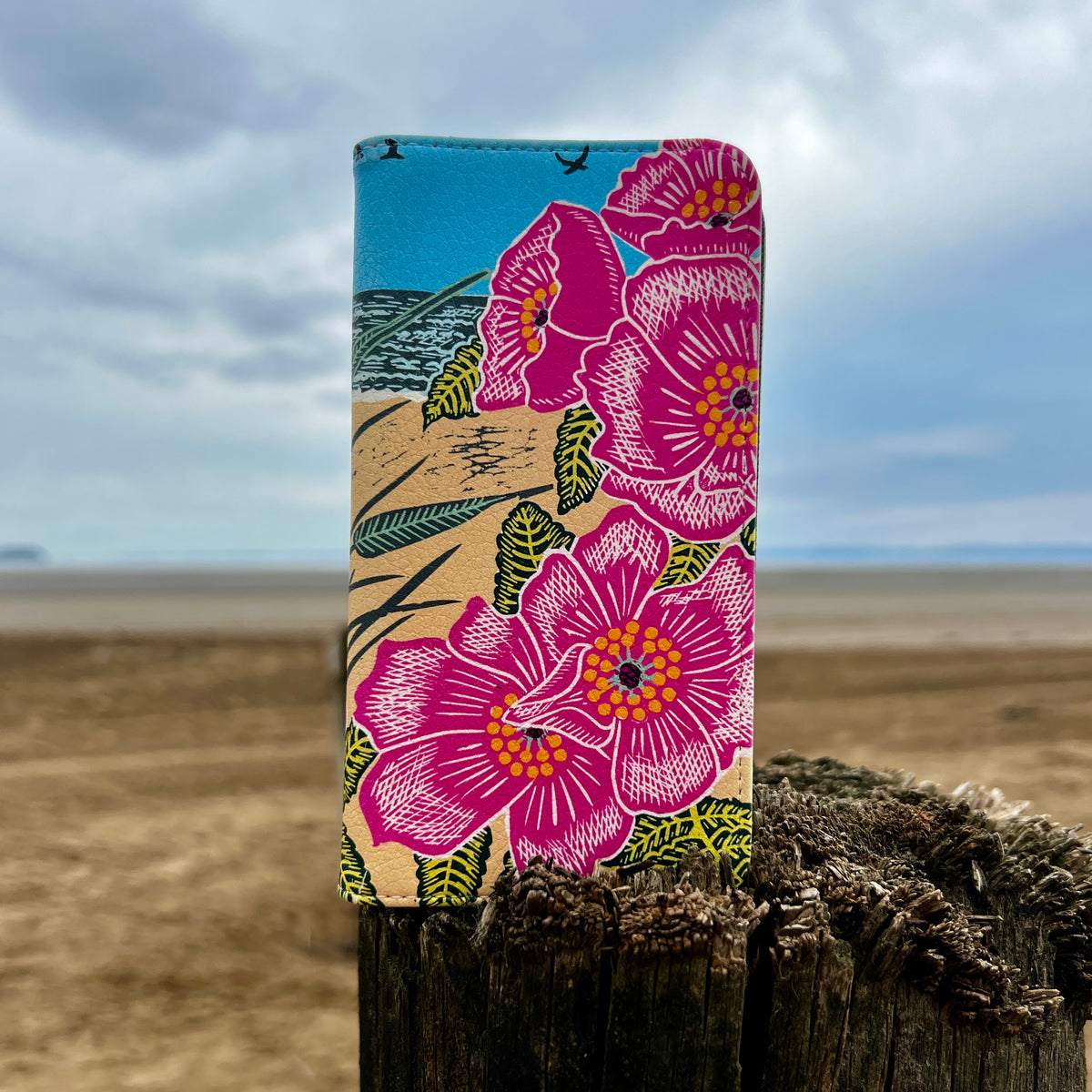 Vegan leather phone folio case featuring a vibrant pink floral Sand Dunes print by Kate Heiss on a wooden post.