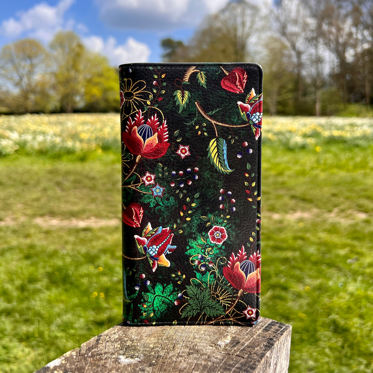 Skelton Green vegan leather folio phone case with a vibrant floral print and satin finish on a wooden post.